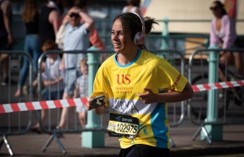 A marathon runner wearing a bright yellow top with Ƶ logo and giant headphones runs along Brighton seafront