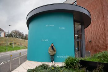 Helen Cammock stands in front of the artwork on an external wall at top of Ƶ Student Centre. The text is painted in white and set against a bright teal background, and reads: whisper  tones  vibrate  foundations
