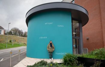 Helen Cammock stands in front of the artwork on an external wall at top of Ƶ Student Centre. The text is painted in white and set against a bright teal background, and reads: whisper  tones  vibrate  foundations